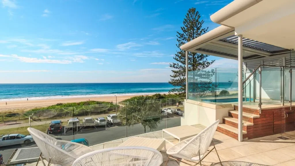 Coastal view from a modern balcony overlooking the beach, featuring a pool, palm trees, and clear blue skies, reflecting the relaxed lifestyle of the Gold Coast.